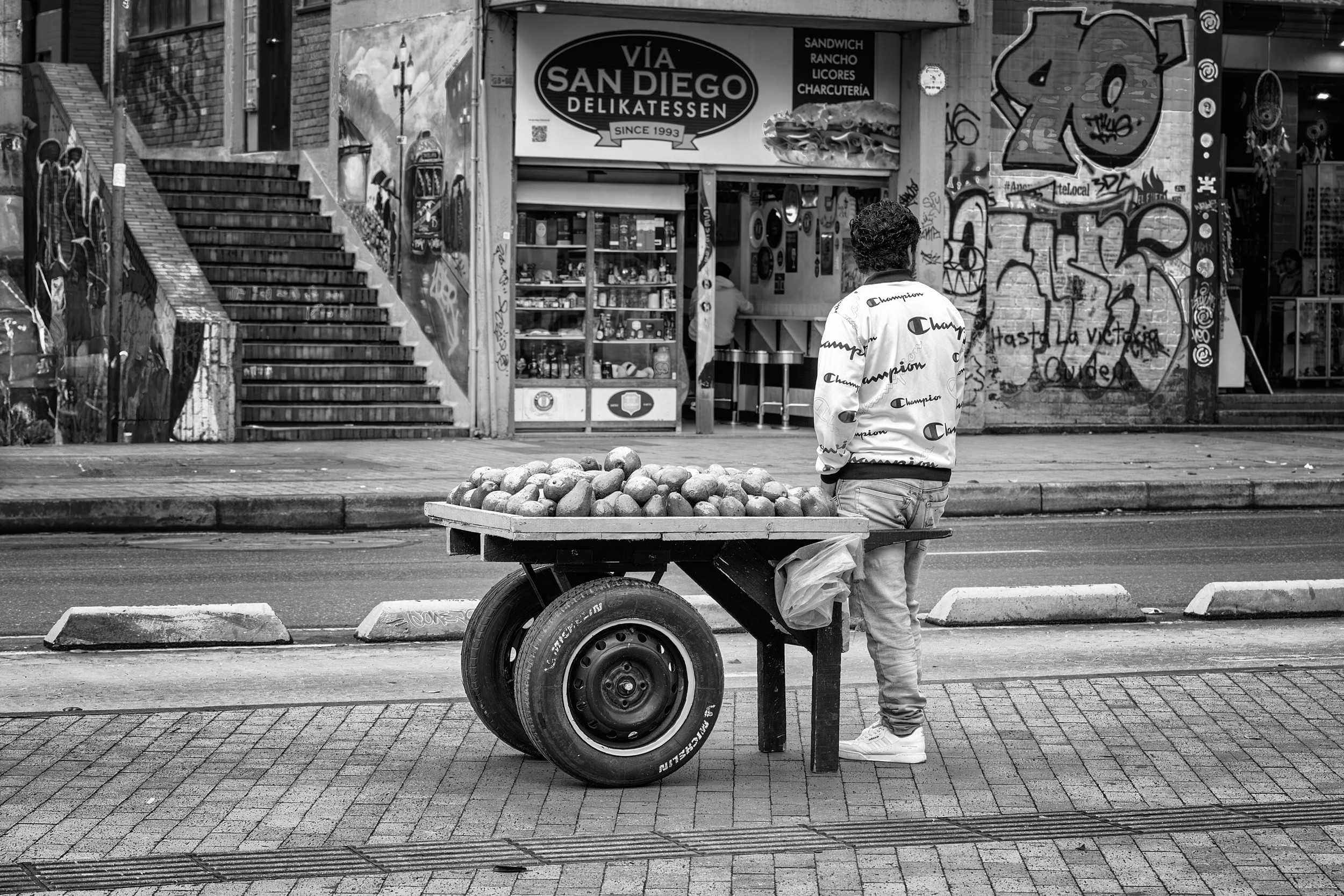 A man stands beside a wooden cart filled with palta (avocados) on a Bogotá sidewalk, facing a deli storefront and graffiti-covered walls.