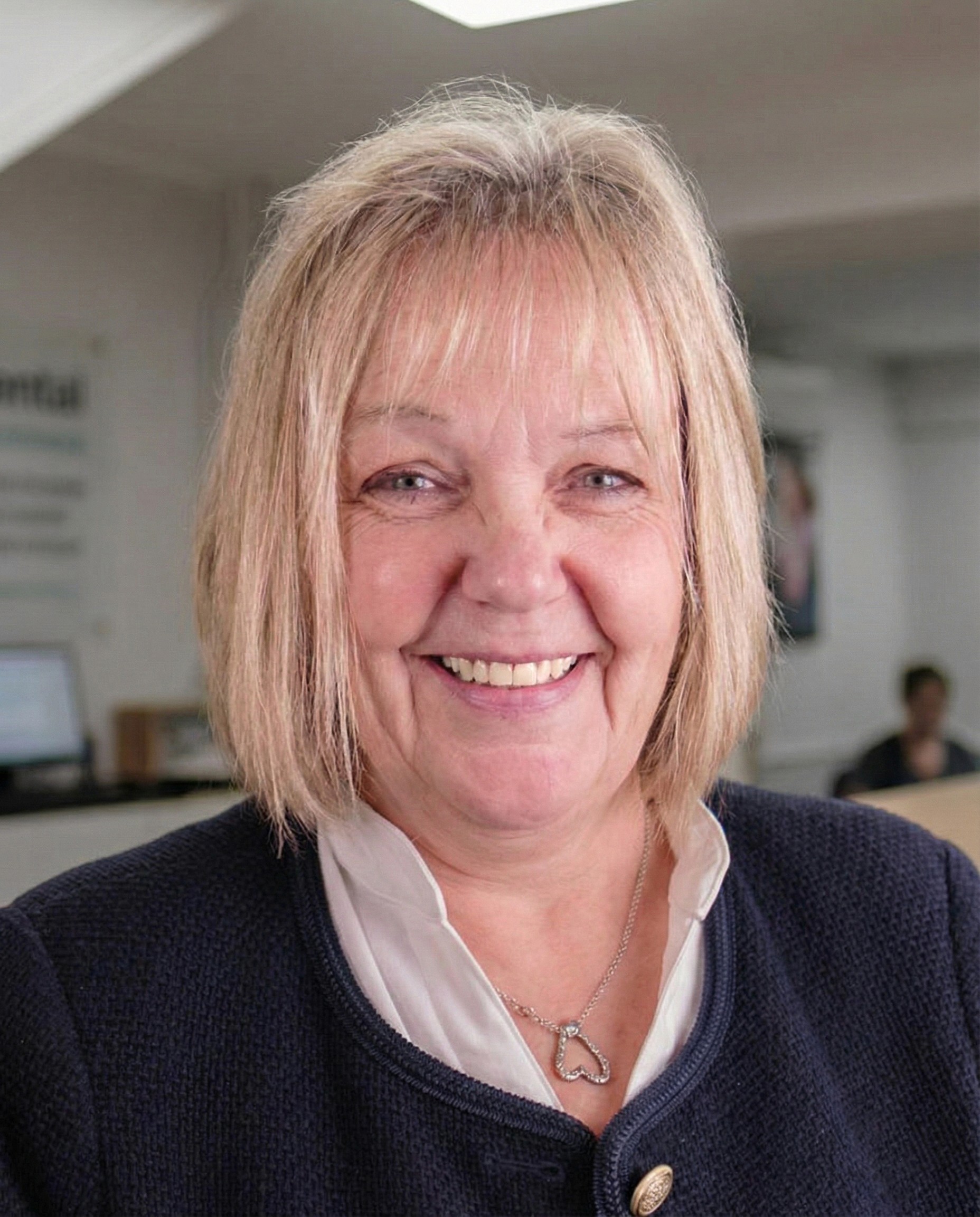 A portrait of Tina, a receptionist at Cricklade Dental Practice, smiling and wearing a white blouse under a dark blue textured jacket with gold buttons. She is wearing a silver heart-shaped necklace and is standing in the practice reception area with the practice logo visible on a glass partition in the background.