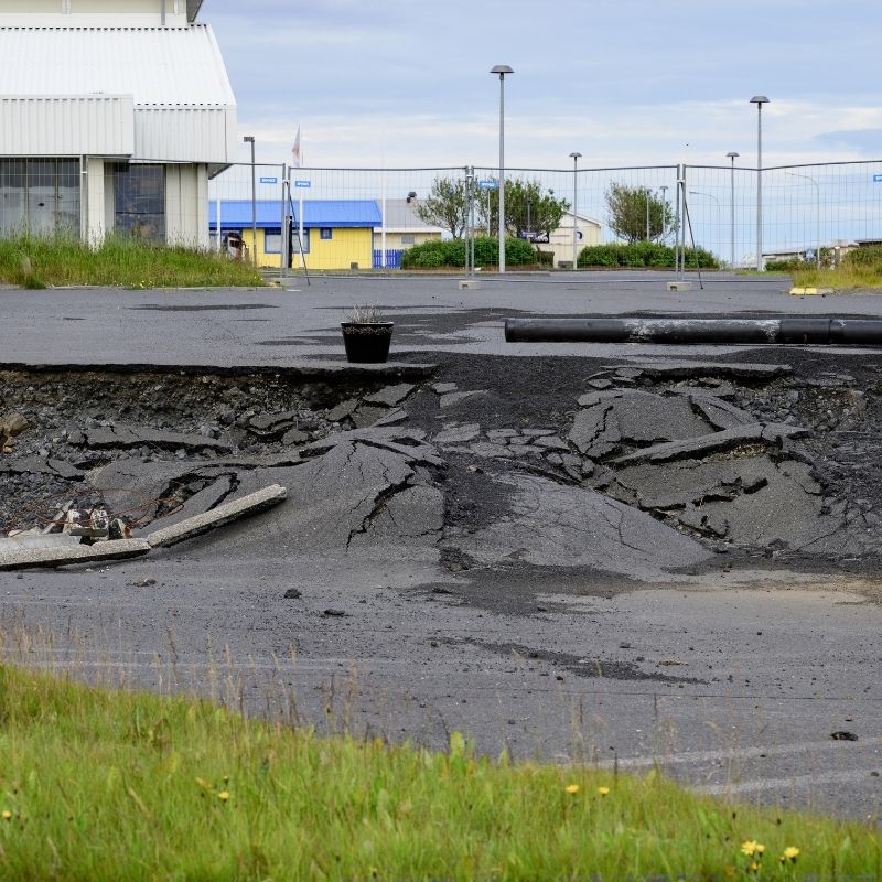 Cracked and collapsed asphalt in Grindavík, Iceland, showing severe ground damage caused by recent earthquake activity, with buildings and fencing visible in the background.