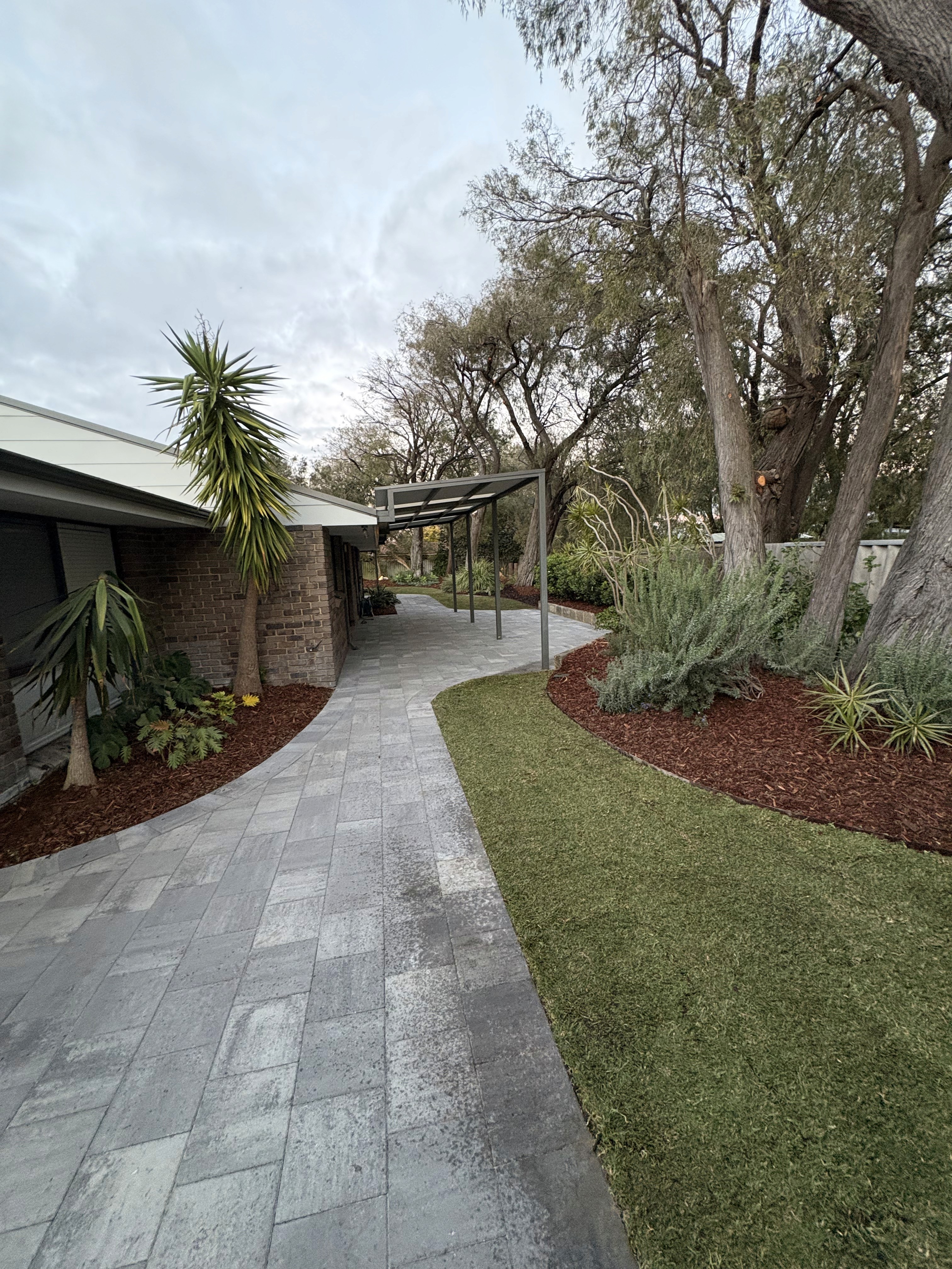 Curved grey paving beside a green lawn, mulched garden beds and a pergola.