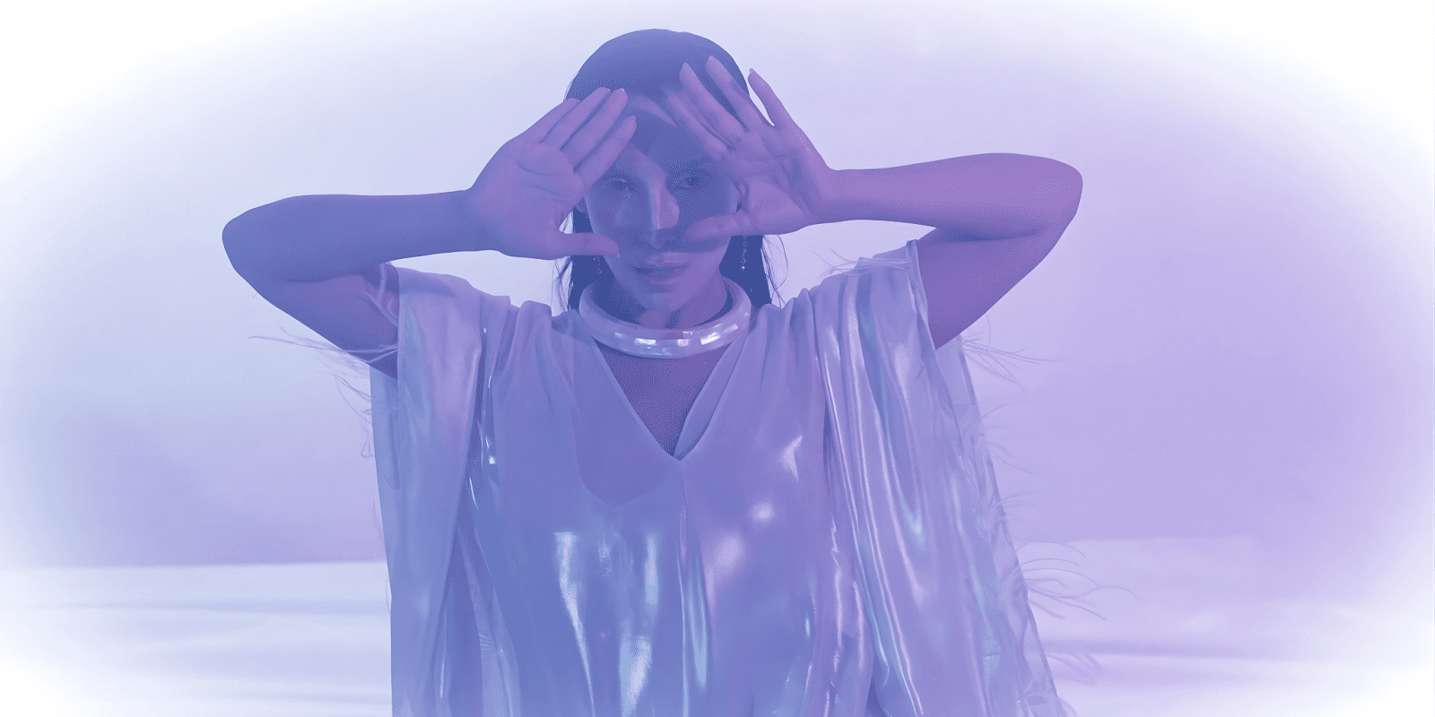 A woman in a metallic silver outfit with a glowing choker, performing with a mystical, ethereal aura in purple light.