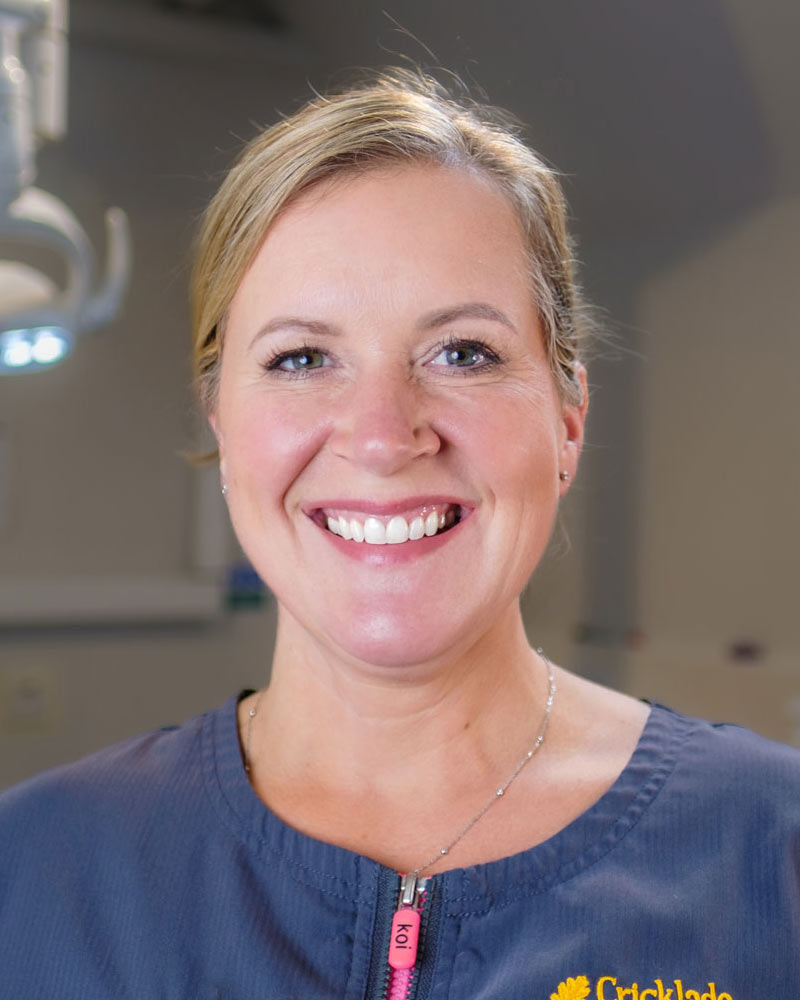 A portrait of Anne, a Dental Hygienist at Cricklade Dental Practice, smiling and wearing a dark grey scrub top with a pink zipper pull and yellow embroidered text that reads "ANNE" on the left and "Cricklade Dental Practice" on the right. She is standing in a dental surgery with a dental light visible in the background.
