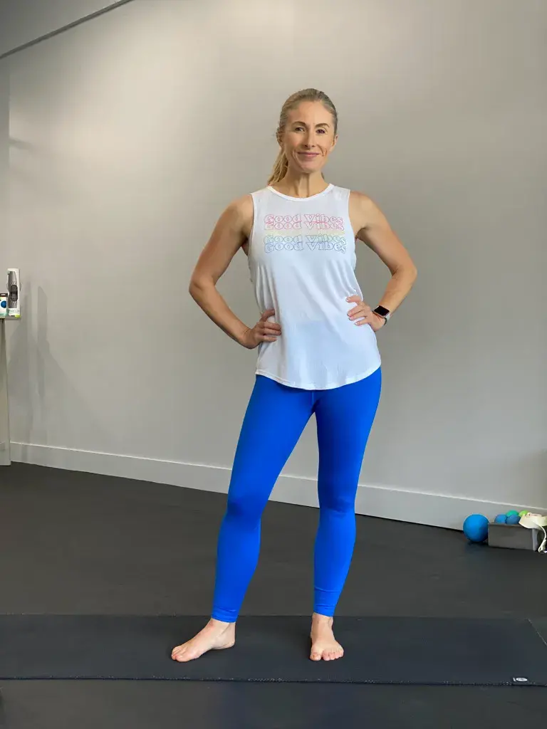 Heather Hausman in a fitness studio stands confidently with hands on hips, wearing a white tank top and bright blue leggings, ready for a workout session.