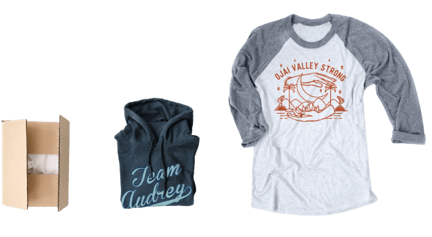 Cardboard box, folded Team Audrey hoodie, and Ojai Valley Strong raglan T-shirt on white background.