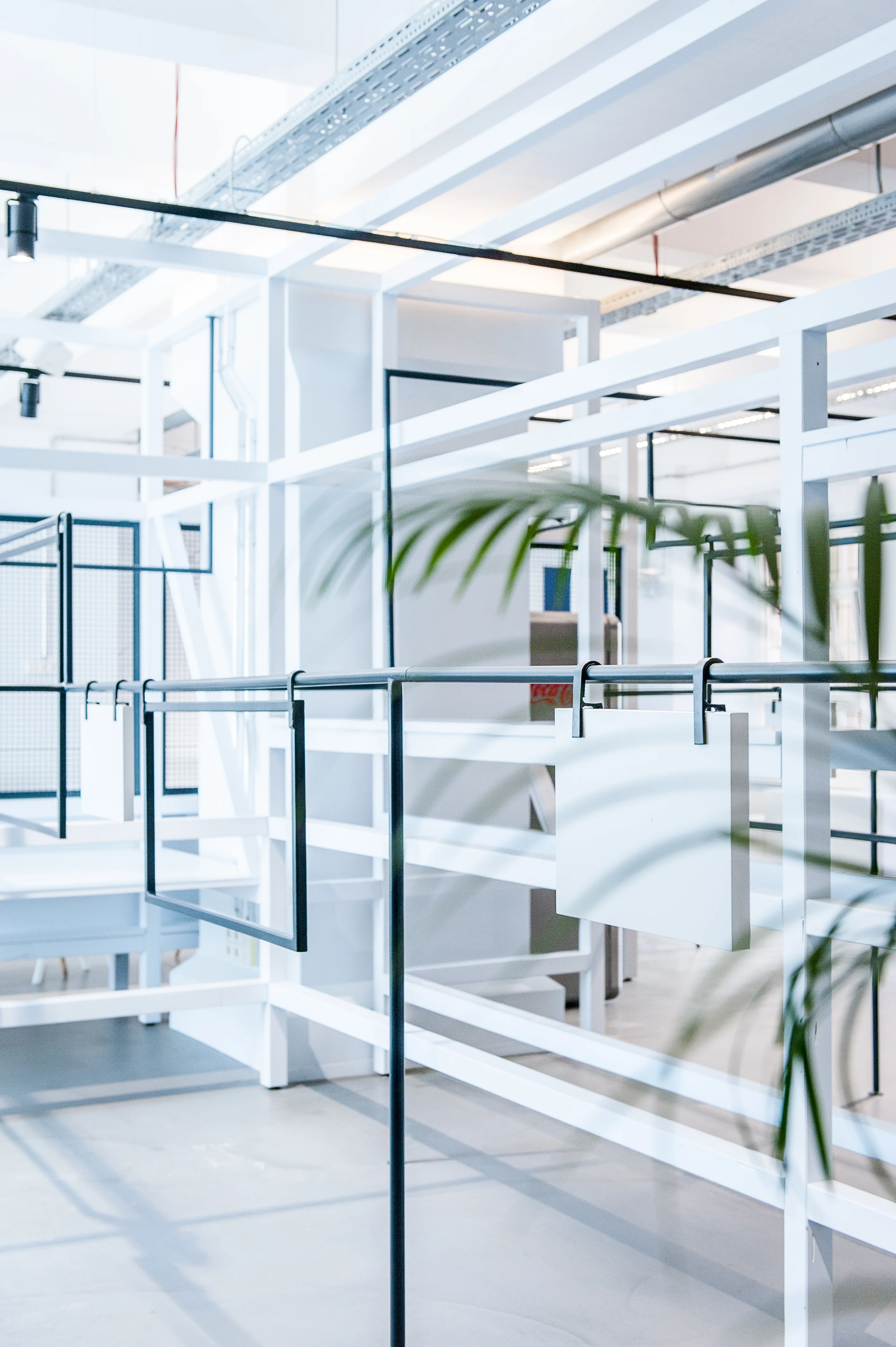 Layered white steel rack structure with black clothes rails and hooks at the 17 FASHION BRANDS showroom, with palm leaves in the foreground.