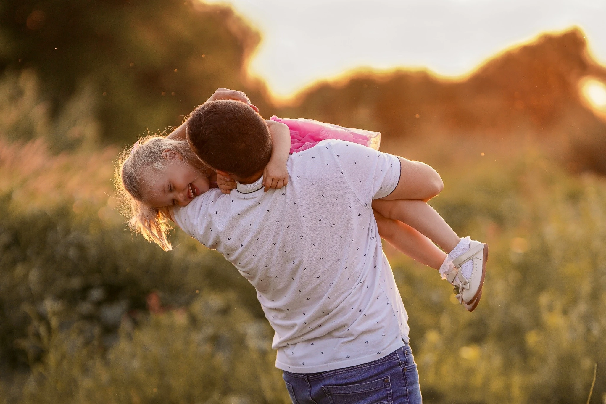  Na zdjęciu widnieje rodzina podczas pikniku na świeżym powietrzu, obok spokojnego jeziora. Składa się z mężczyzny, kobiety i małego dziecka. Mężczyzna ma na sobie jasnoniebieską koszulę i krótkie spodenki, siedzi z przodu i trzyma dziecko na kolanach. Kobieta, ubrana w kolorową sukienkę i kapelusz, siedzi obok i uśmiecha się, patrząc na dziecko. Sceneria jest zielona, z dużą ilością trawy i drzew w tle. Całość tworzy spokojną i sielankową atmosferę.