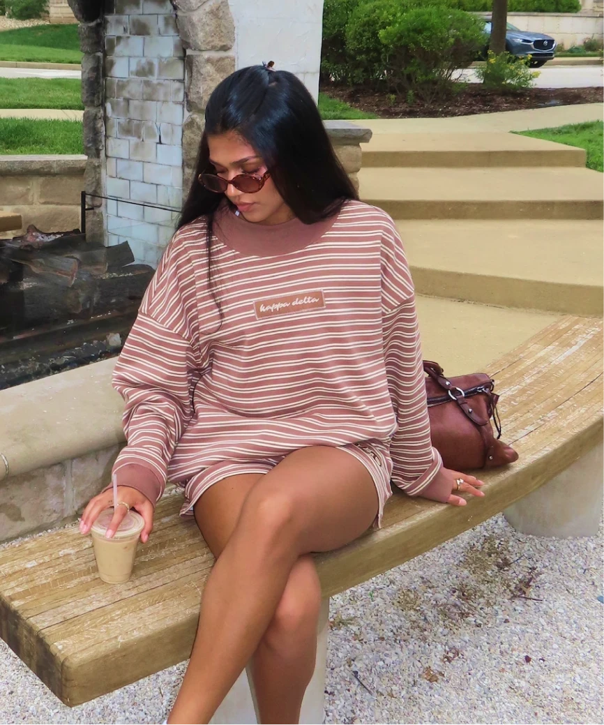 Sorority girl Meggie Eyer in a brown striped Kappa Delta sweatshirt, sunglasses on, sitting on a bench with an iced coffee.