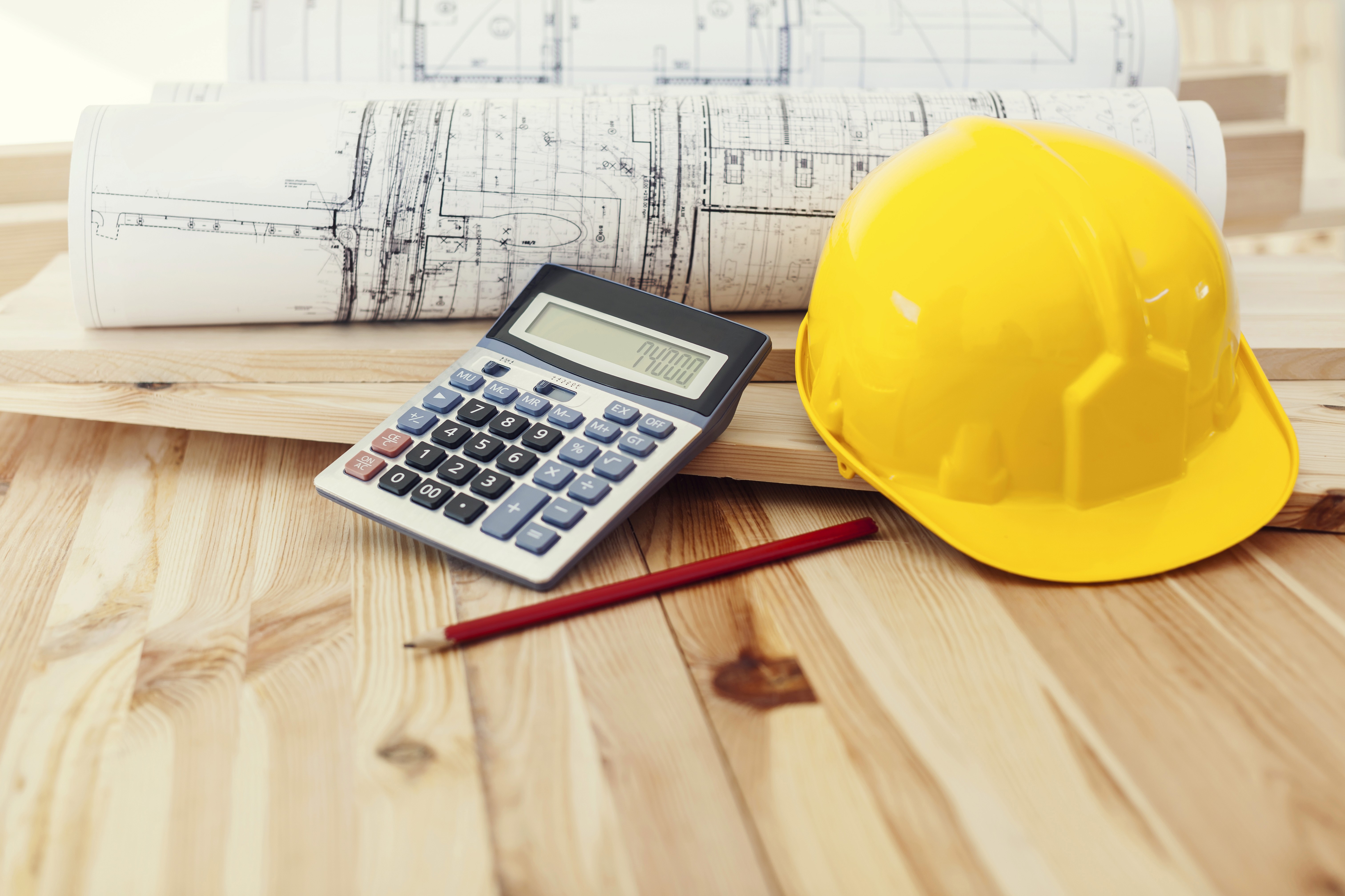 Yellow hard hat and calculator on wooden table with rolled-up blueprints and red pencil, suggesting a construction and load calculation planning scene.