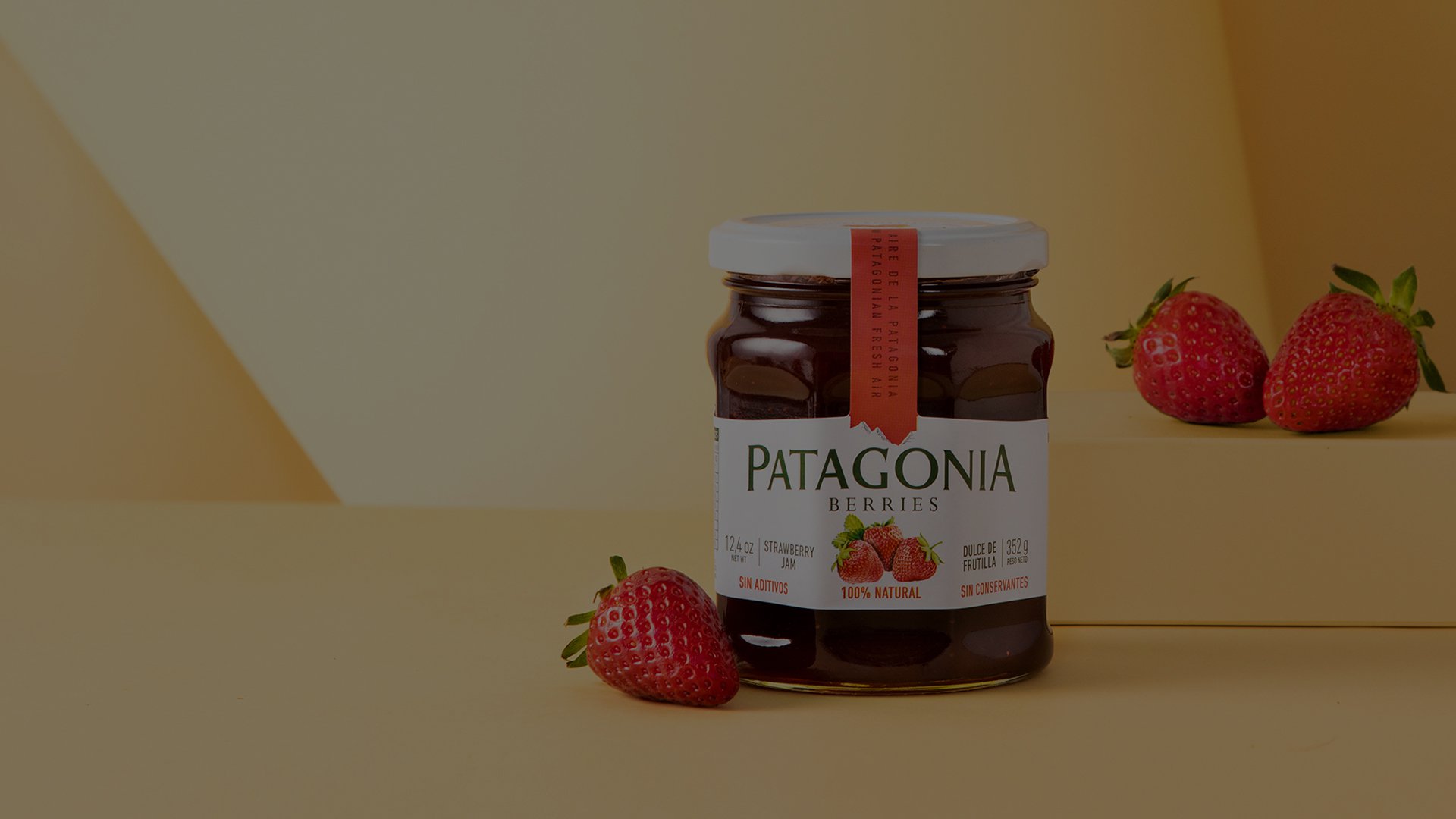 A jar of Patagonia Berries strawberry jam on a yellow surface, accompanied by fresh strawberries. The scene is warm and inviting with soft lighting.