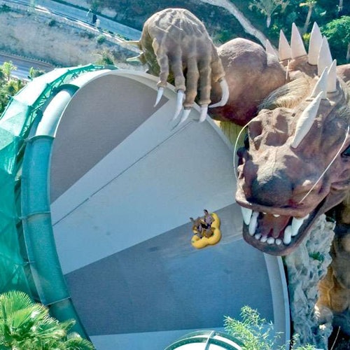 People in a yellow raft descending a large water slide shaped like a dinosaur's mouth, with surrounding greenery and a pathway.