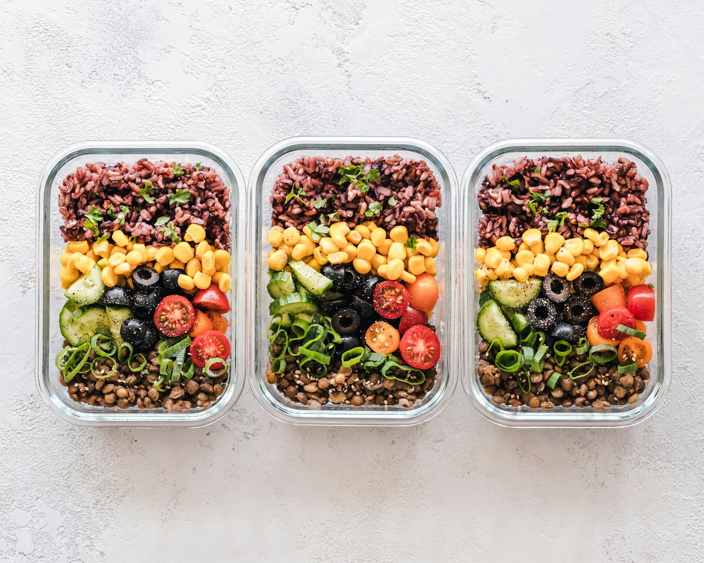 Three meal-prep containers filled with rice, corn, tomatoes, cucumber and lentils, showing a balanced weekly fueling plan for runners