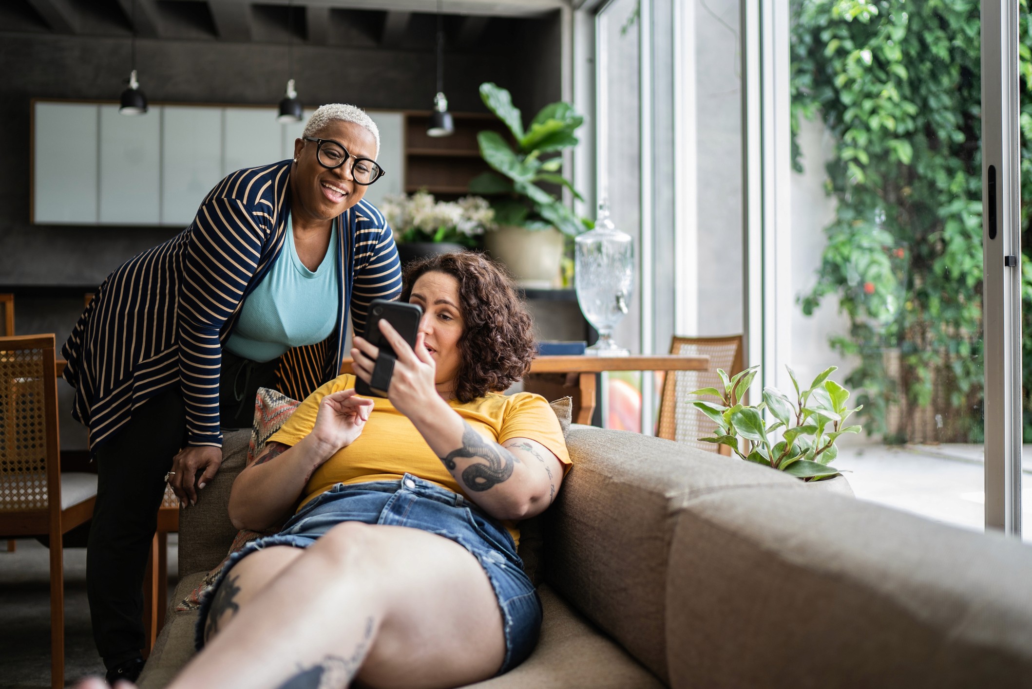 A woman sitting with her legs up on a couch on her phone, conversing with another women who is leaning aginist the couch also looking at the phone