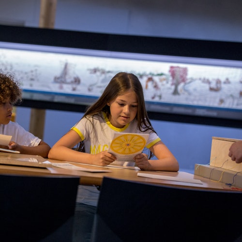 孩子们坐在桌子旁，研究教育材料。背景中可以看到一个展示绘画或艺术作品的大屏幕。