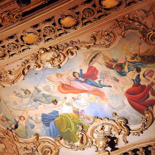 Ornate ceiling mural with figures in colorful clothing engaged in joyous activities, surrounded by intricate gold decorations.