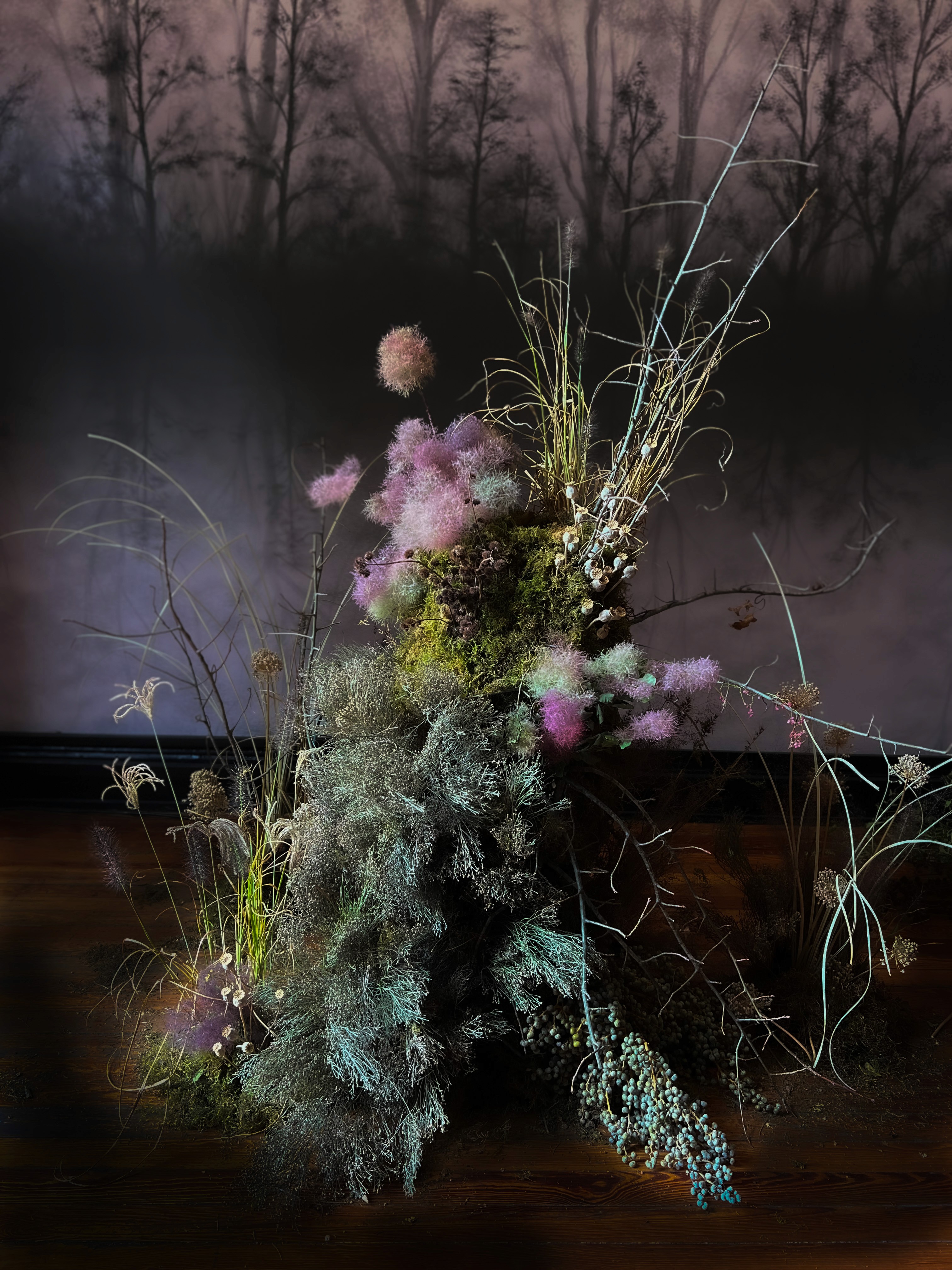 Instalación floral artística con ramas y flores etéreas en tonos lilas y verdes sobre fondo oscuro, de estilo escenográfico y poético.