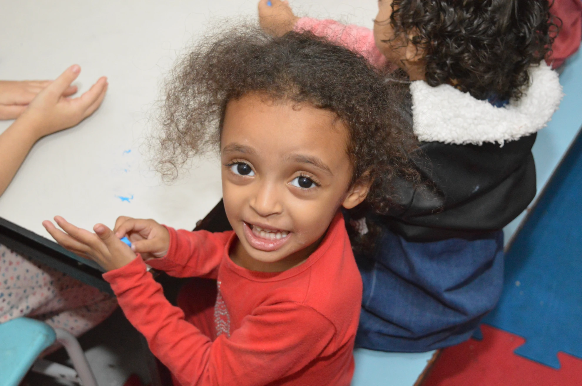 Criança sorrindo e mostrando um trabalho de papel em mesa de atividades com outras crianças ao redor.