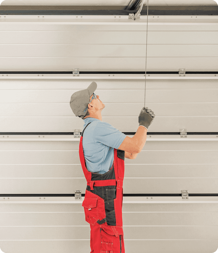 Professional technician in red overalls performing garage door service on a sectional residential door. Close-up of expert technician adjusting the emergency release cord and track alignment for secure and reliable operation.