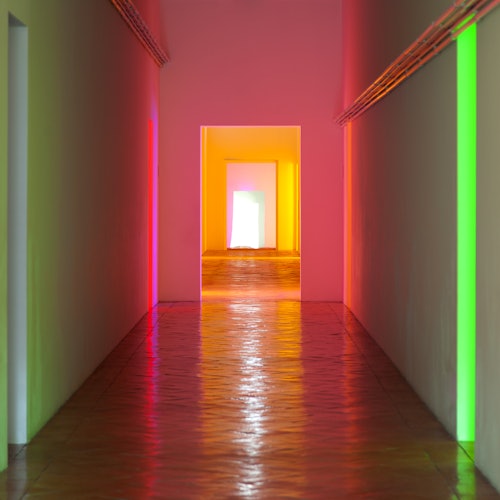 En hall med färgglad belysning i rosa, grönt och gult som reflekteras på väggar och golv, som leder till en öppen dörröppning.