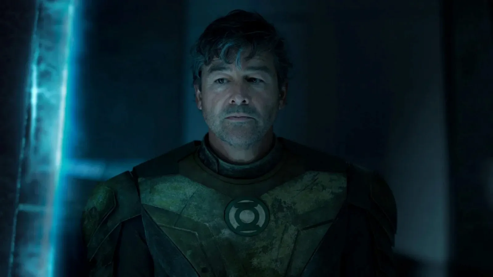 A person in a dimly lit setting wears a weathered green military-style armour, featuring a round emblem on the chest, and stands pensively in front of a metallic blue-tinted background.
