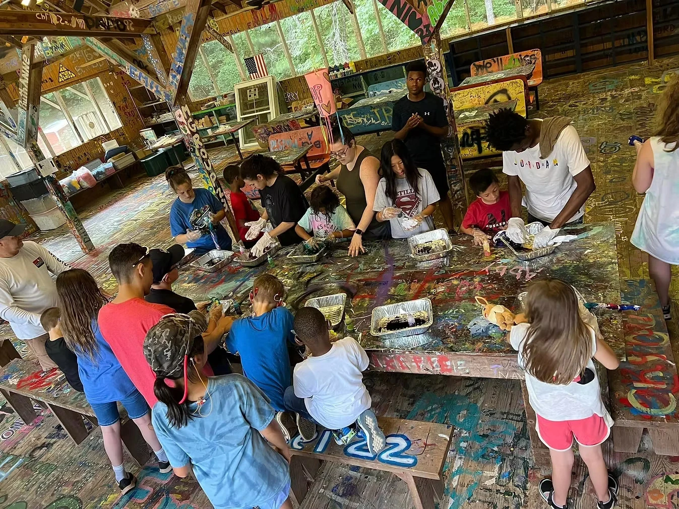 A large group of campers and staff participate in a craft activity at Camp Juliena. They are sitting around a long table that is covered in paint.