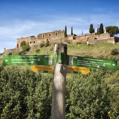 Tanda ng kahoy na may berdeng at dilaw na mga direksyon sa harap ng isang lumang batong kuta sa isang damuhang burol sa ilalim ng asul na langit.