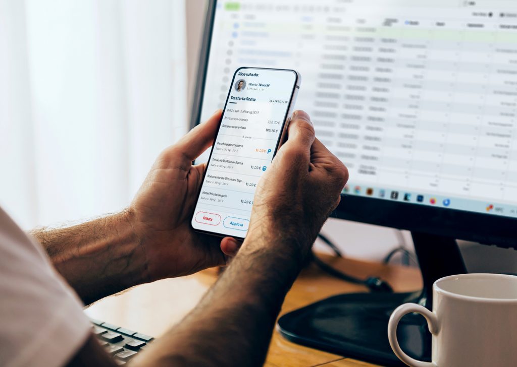 Una persona confronta la nota spese inserita sul software gestione spese dello smartphone con i dati integrati sullo schermo del pc