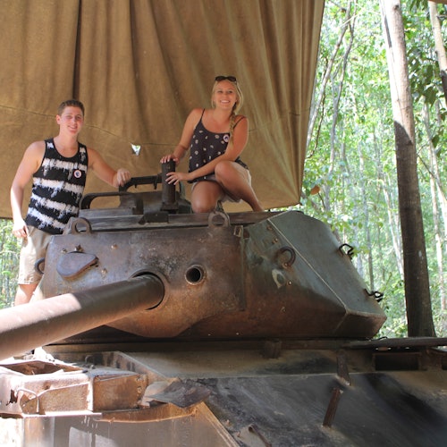 Dua orang berpose di atas tank tua di bawah kanopi dalam latar hutan, dengan pepohonan terlihat di latar belakang.