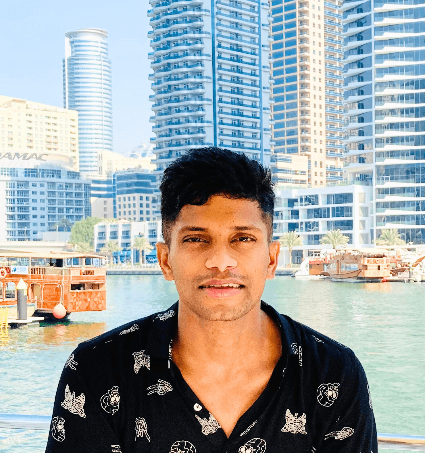 A young person with curly hair smiles at the camera while sitting in front of a laptop against a dark background.