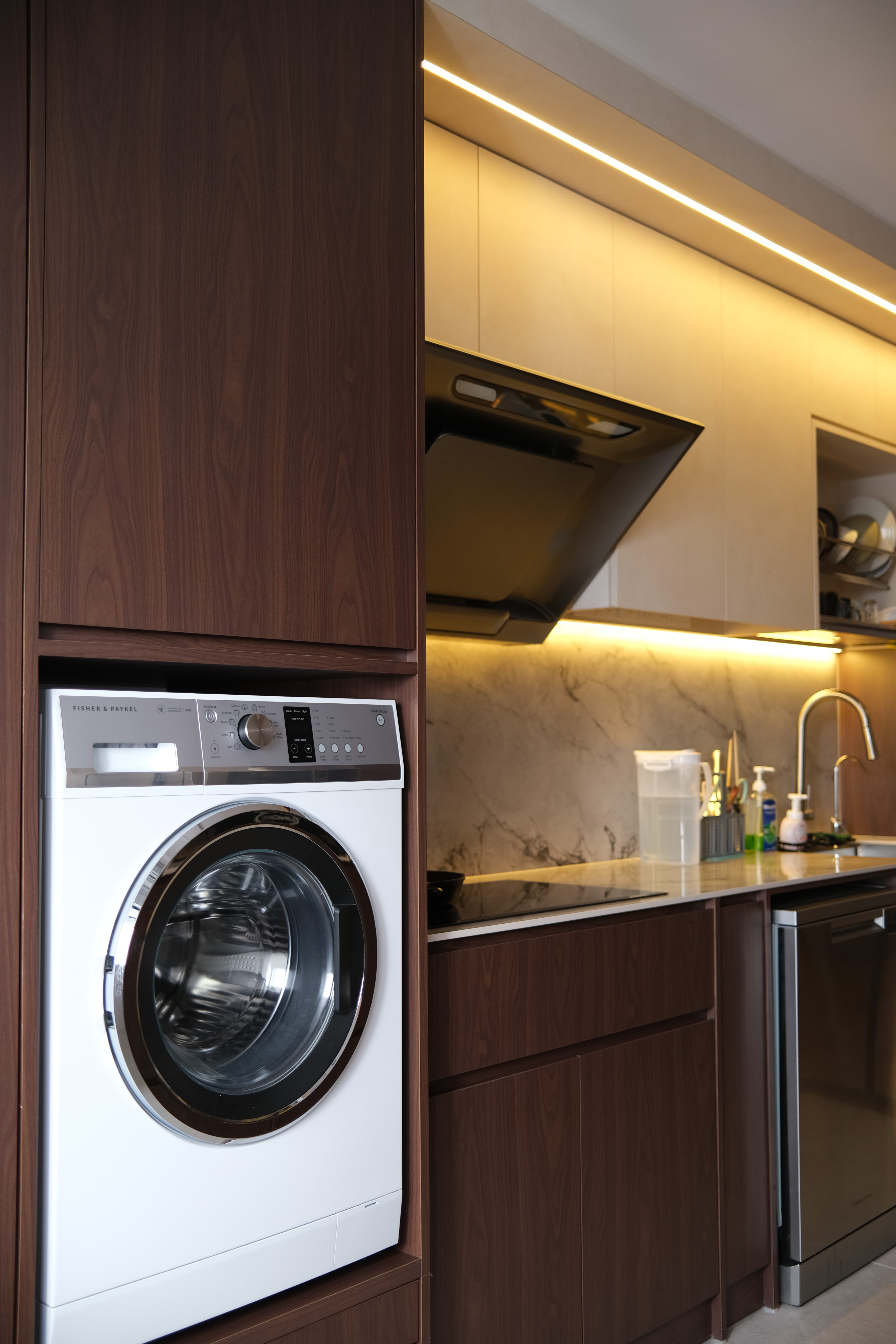 A seamlessly integrated kitchen and laundry zone where warm timber cabinetry, stone surfaces, and concealed appliances prioritise efficiency without compromising visual clarity.