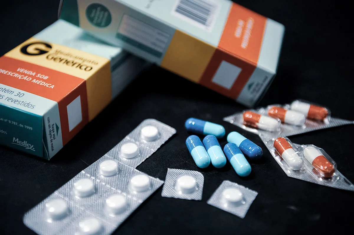 Caixas de medicamentos genéricos com tarja vermelha ao lado de cartelas de comprimidos brancos e cápsulas coloridas espalhadas sobre fundo escuro.