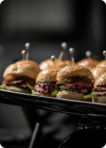 Mini hambúrgueres gourmet servidos em bandeja preta para evento corporativo premium, com apresentação sofisticada e iluminação intimista.