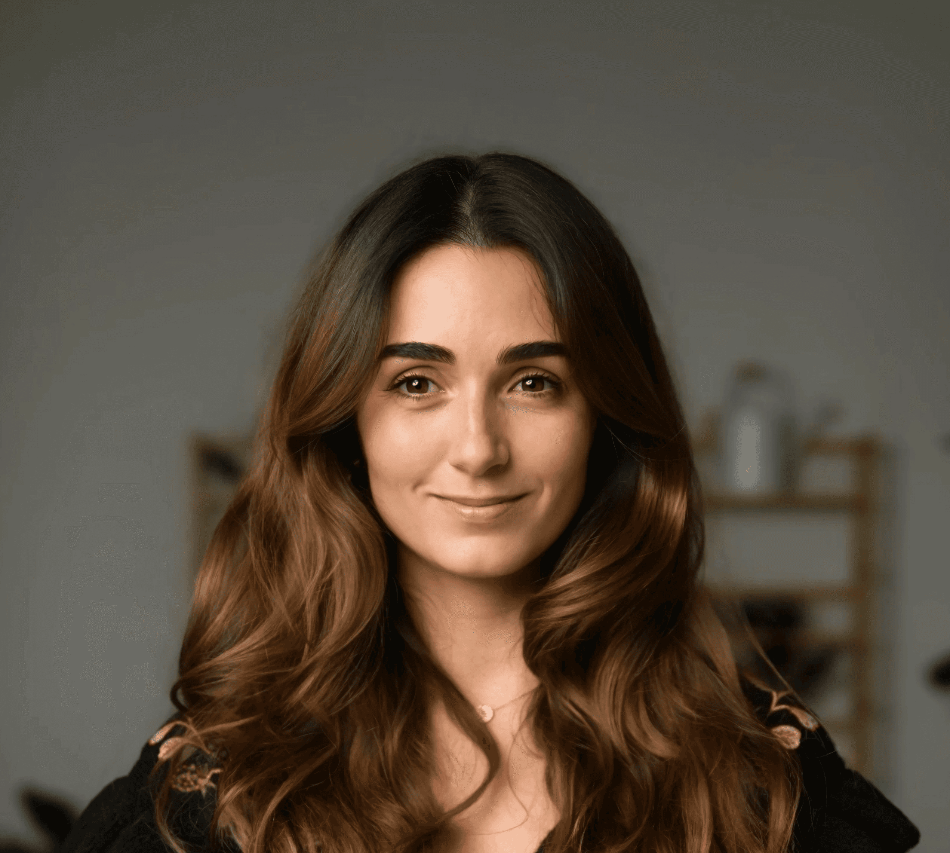 A smiling woman with long, wavy brown hair, wearing a dark top against a soft gray background representing an author for.