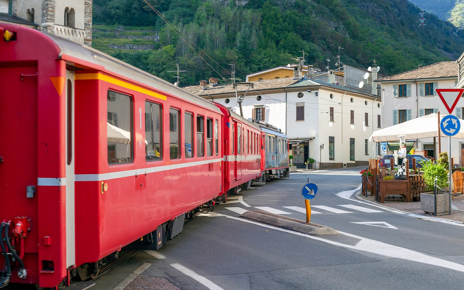 Bernina treni, arka planda binalar ve dağlarla birlikte İtalya'nın Tirano şehir caddesinden geçiyor.