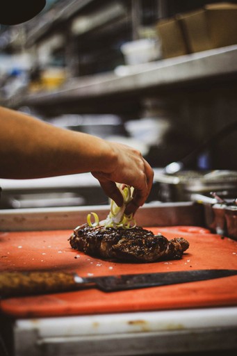 Close-up of a chef's hand placing sliced scallions on a grilled steak. The steak rests on an orange cutting board.