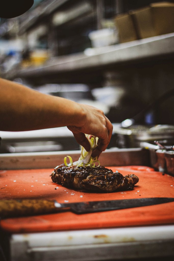 Close-up of a chef's hand placing sliced scallions on a grilled steak. The steak rests on an orange cutting board.