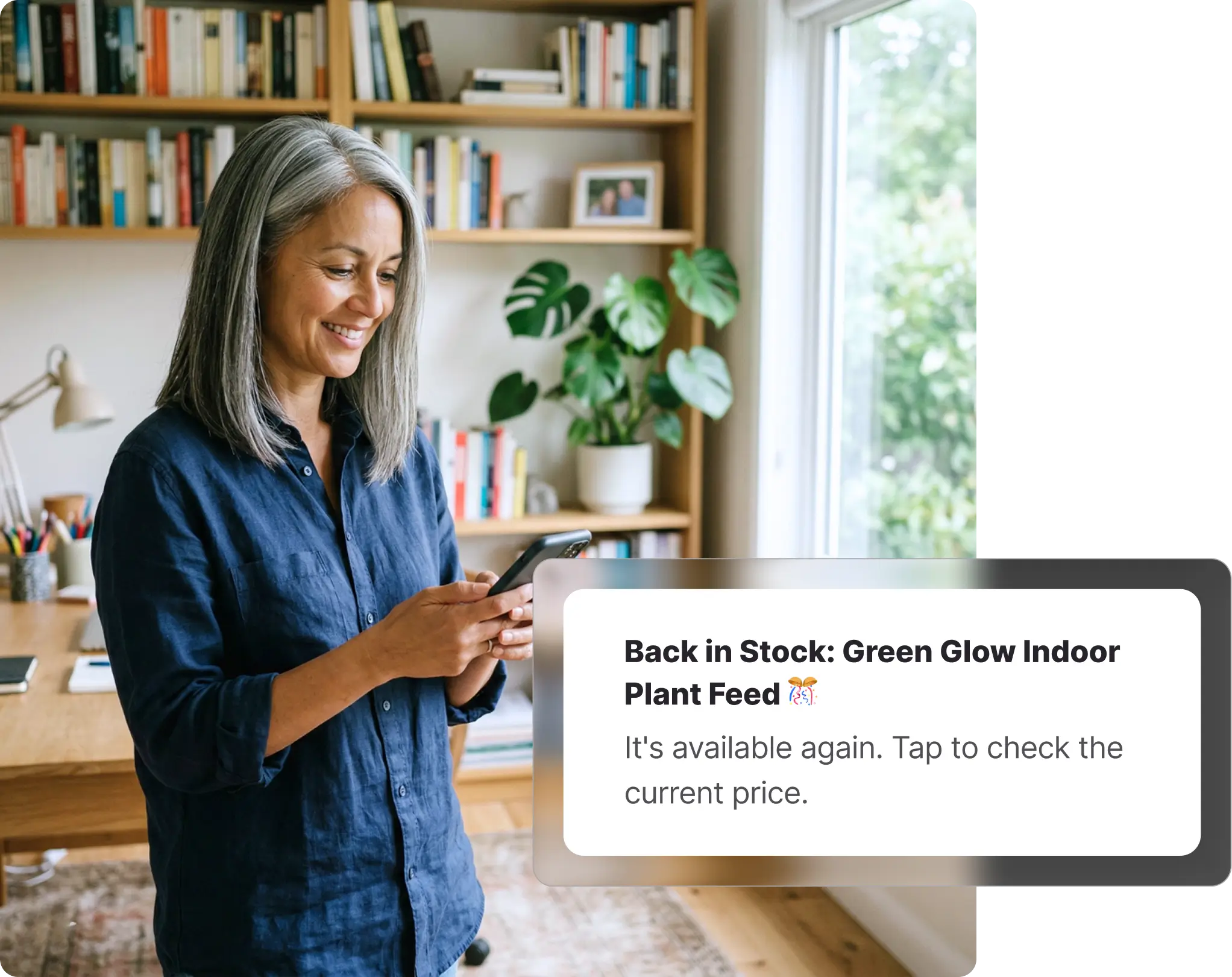 Older woman with silver hair standing in a bright home office checking her phone, receiving an Amazon back-in-stock notification for plant feed on Whisprice, with bookshelves and indoor plants visible behind her.