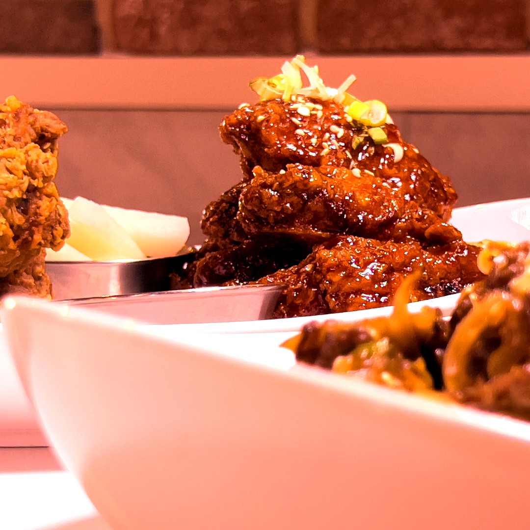 Koreanz platter featuring savory bulgogi in the foreground, accompanied by crispy fried chicken and sticky Yangnyeom chicken in the background. Served with pickled radish and traditional banchan, this dish highlights the rich, authentic flavors of Korean BBQ and comfort food.