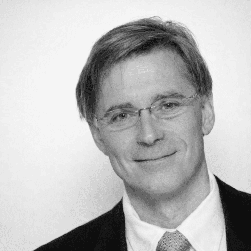 A person with medium-length brown hair wearing a dark, textured coat and a burgundy turtleneck. They have a calm, neutral expression.