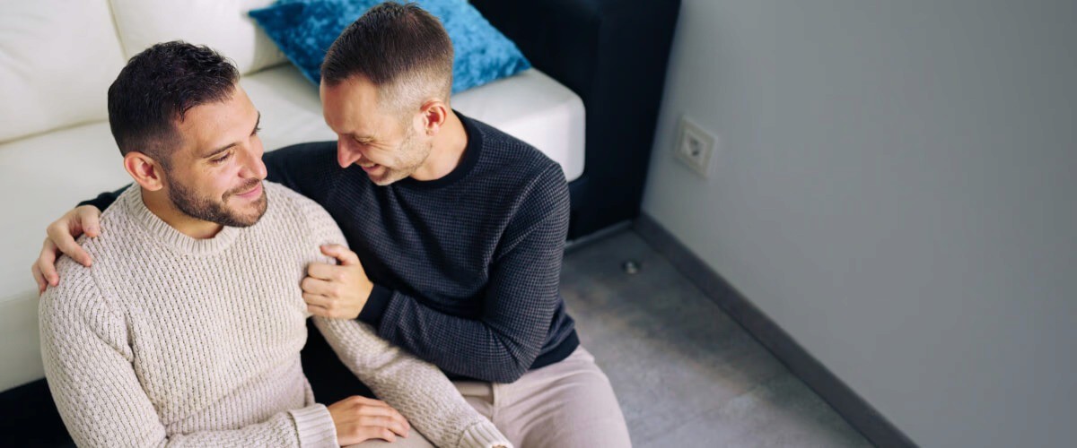 Two men smiling and embracing on floor next to sofa. One wears cream sweater, the other black.