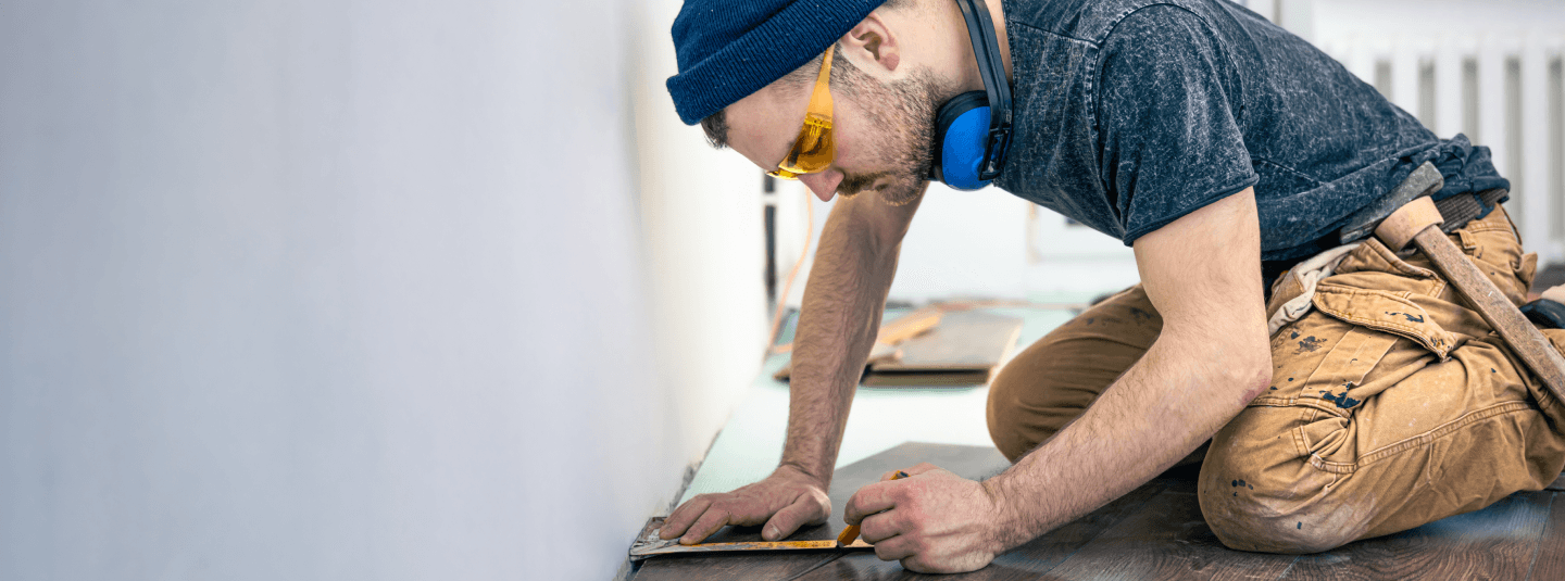 A man wearing a beanie, protective glasses, and headphones kneeling on the floor, carefully measuring or working on flooring installation in a well-lit indoor space.