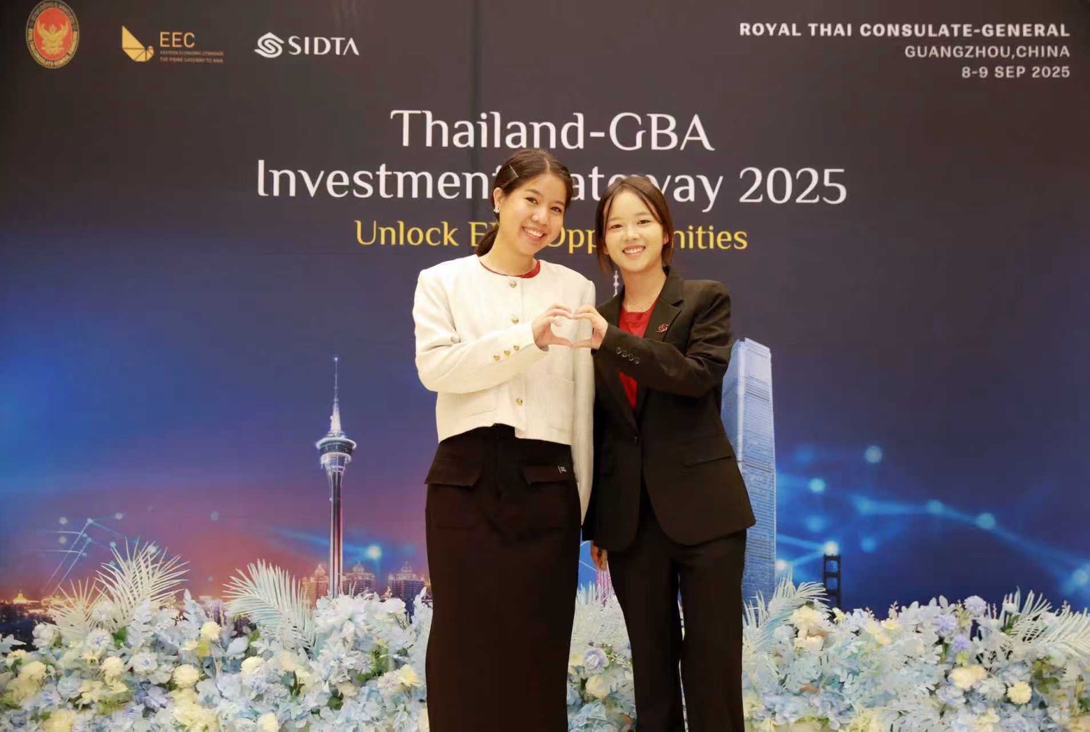 Emily Praewa and Bin Wang from the Thailand–GBA Investment Gateway 2025 making a heart shape with their hands in front of the event backdrop in Guangzhou, China — capturing the spirit of teamwork and friendship behind the scenes.