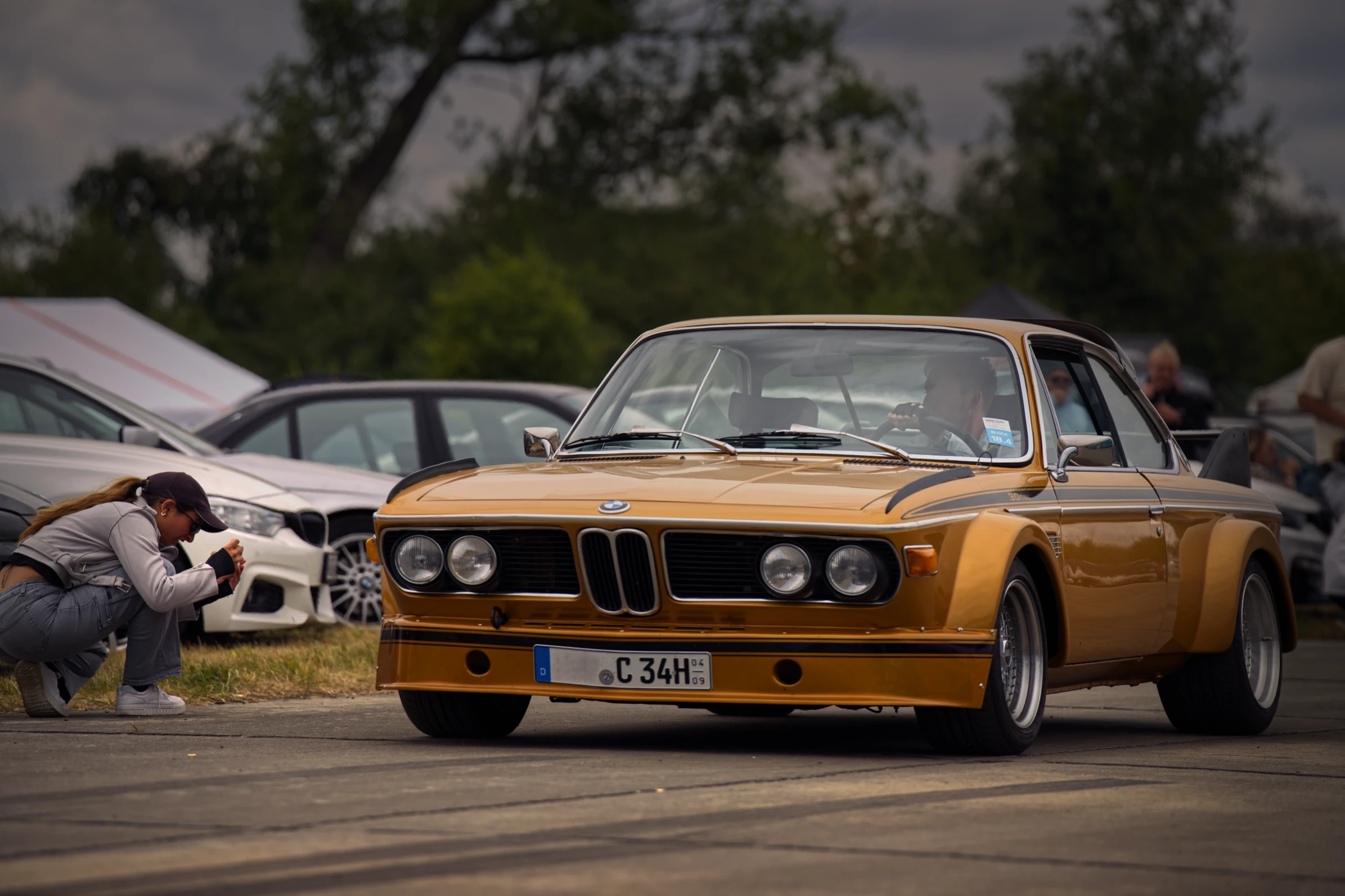 Fotografin vor einem goldenen BMW E9 CSL beim BMW Syndikat Asphaltfieber 2025 – Begegnung von Automobilkultur und Fotografie.