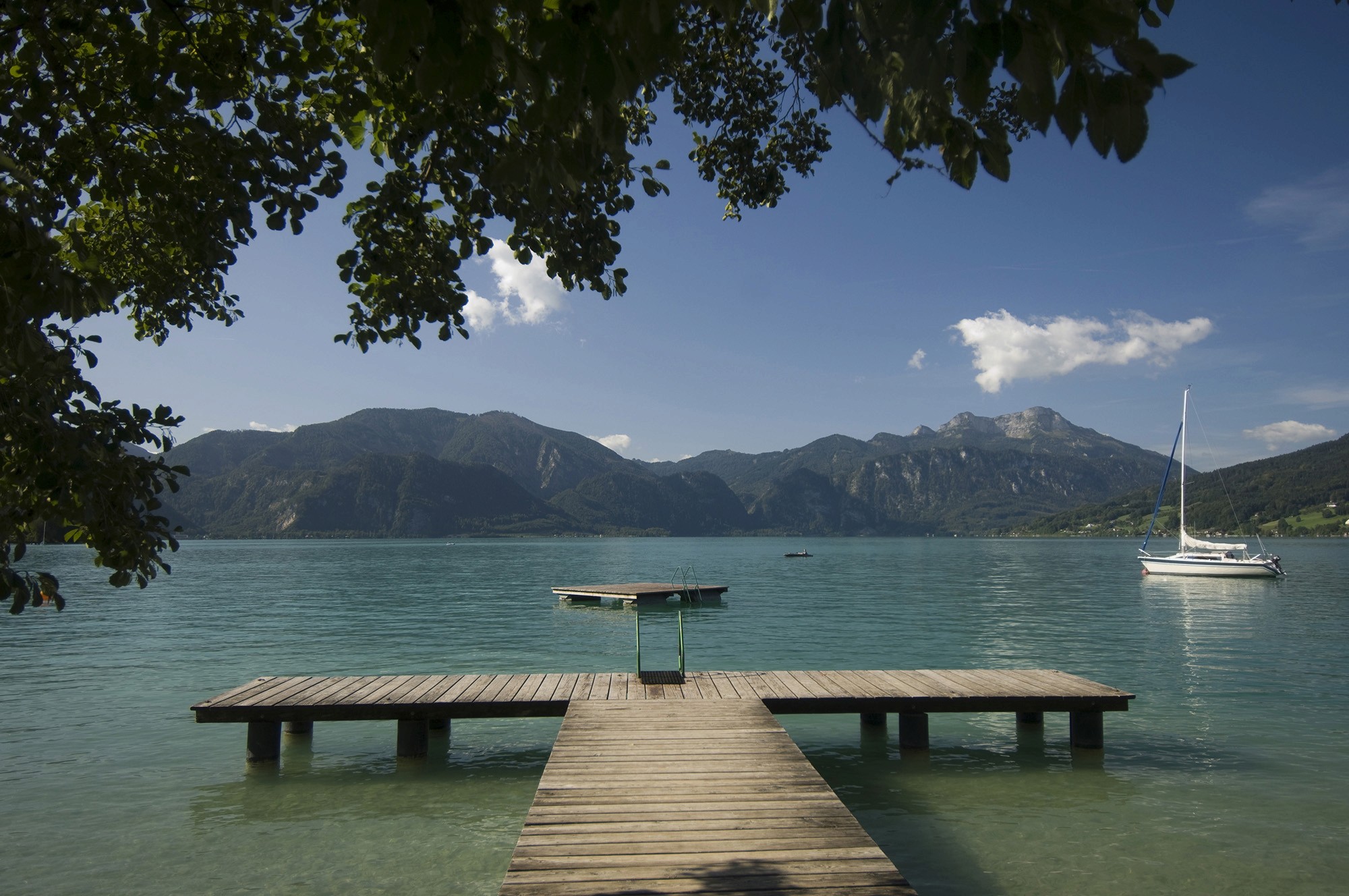 Holzsteg am Attersee in mit Bergen im Hintergrund und einem Segelboot auf dem Wasser.