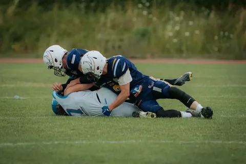 Two football players in blue uniforms tackle an opposing player in a white uniform on a grassy field during an intense game.