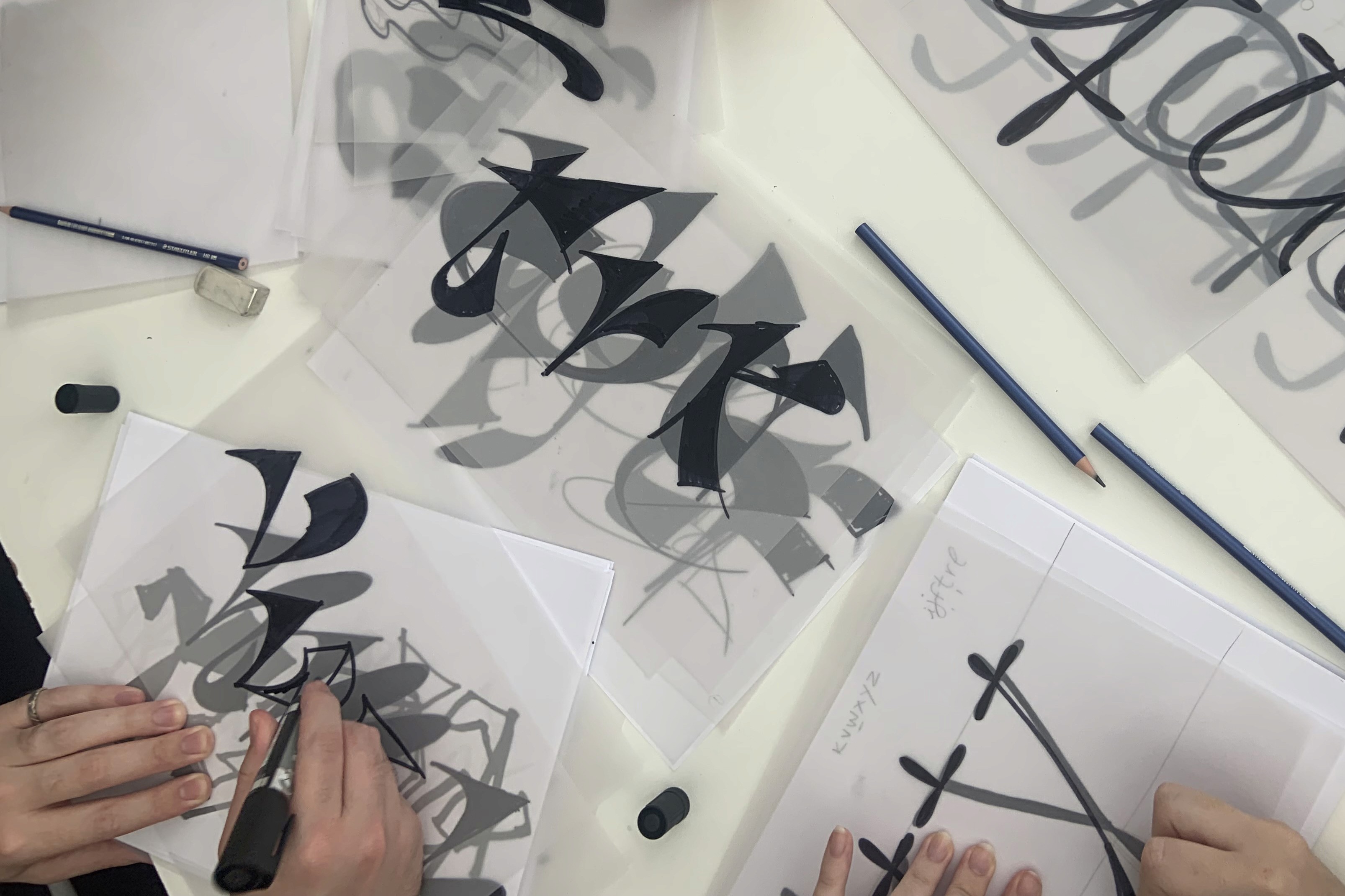Overhead view of hands drawing black letterforms on transparent sheets at a workshop table.