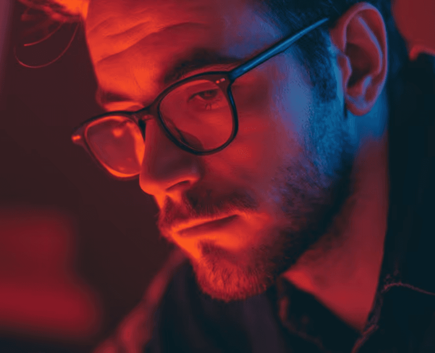 A man wearing glasses is illuminated by red and blue lighting, creating a moody and introspective atmosphere.
