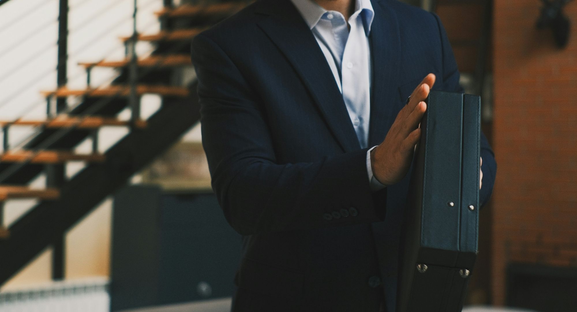 imagem cortada (rosto e parte inferior) de um homem vestido de terno azul escuro segurando uma mala