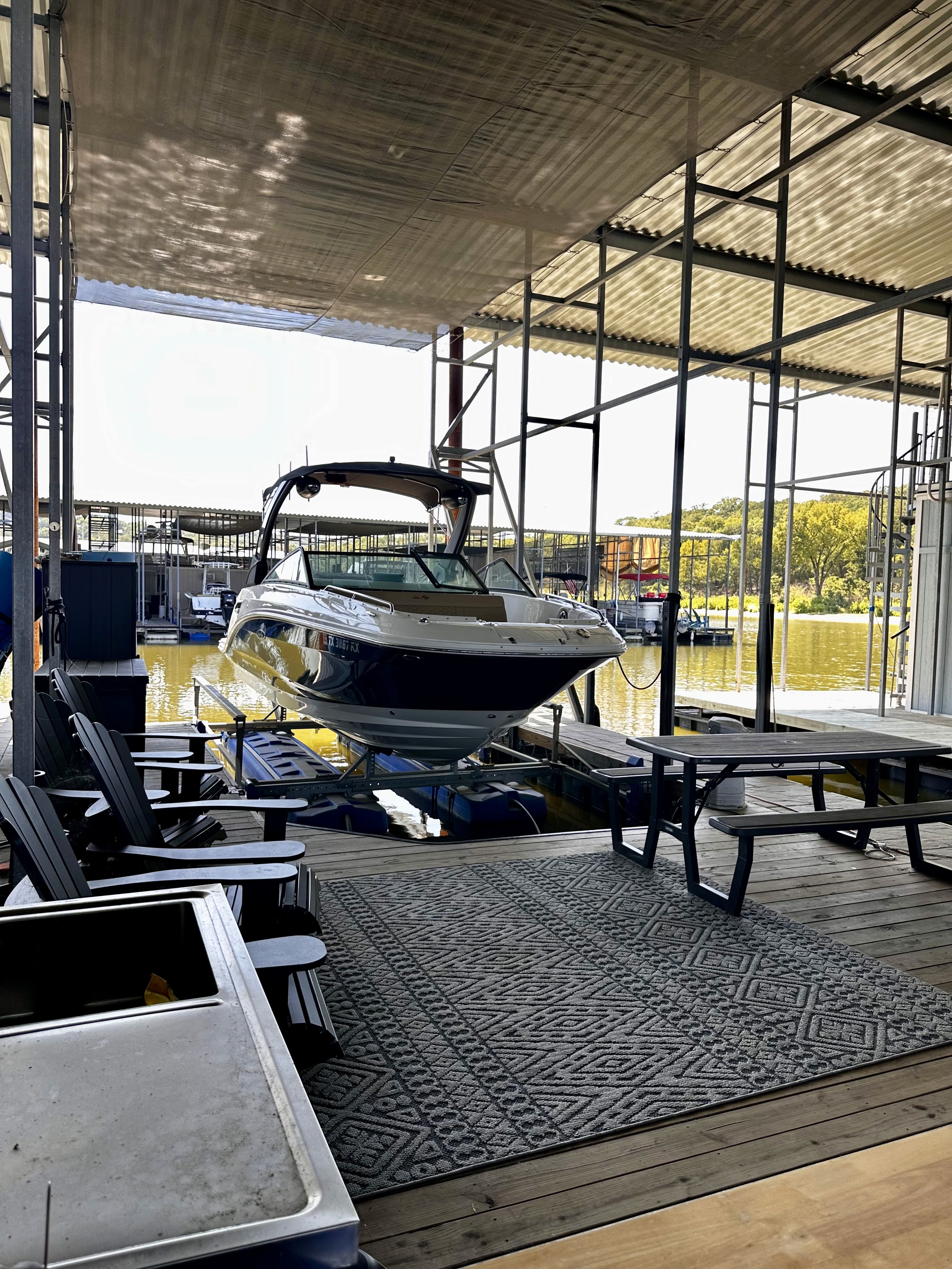 A sleek speedboat is docked on a covered marina slip, surrounded by outdoor seating and tables, with a sunlit view of the water beyond, emphasizing leisure and water activities.