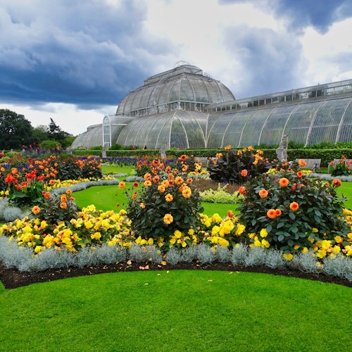A large glass conservatory behind a vibrant garden with colorful flowers and neatly manicured green lawns under a cloudy sky.