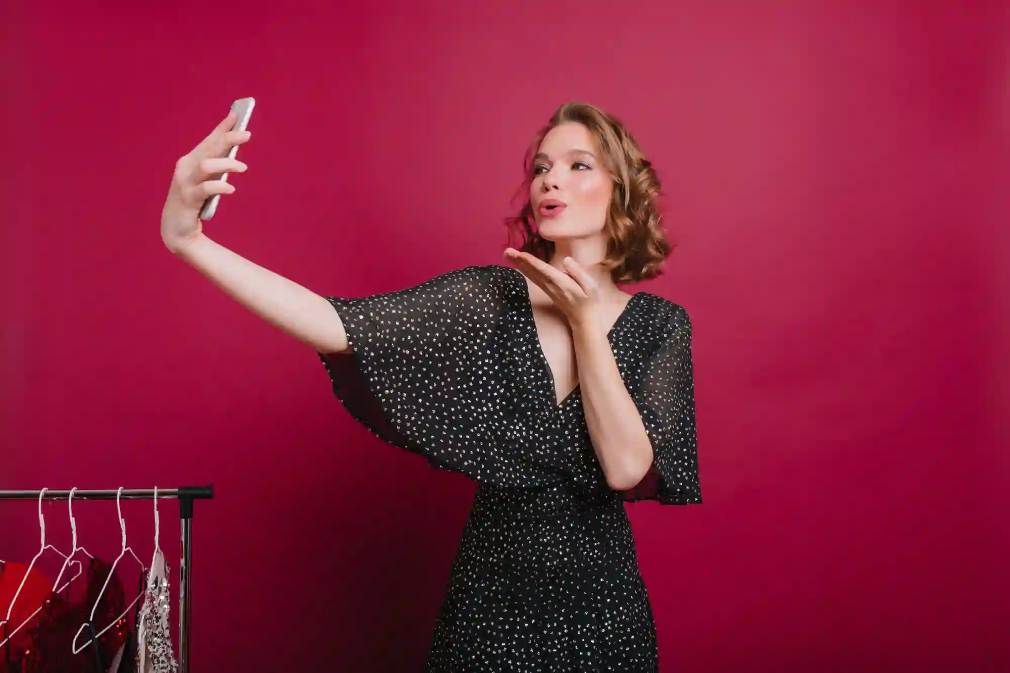 A woman in a sparkly black dress blows a kiss while taking a selfie against a deep pink background.